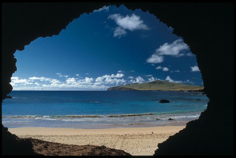 Osterinsel - Blick von einer dunklen Höhle auf einen Sandstrand, das Meer und eine bewachsene Insel unter blauem Himmel mit Wolken