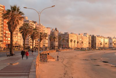 Promenade mit Palmen und Menschen neben einem breiten Sandstrand vor einer langen Reihe von Wohngebäuden bei Sonnenuntergang