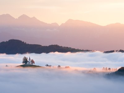 Kirche auf einem Hügel ragt aus Nebelmeer mit Gebirge im Hintergrund beim Sonnenaufgang