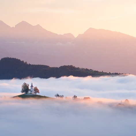 Kirche auf einem Hügel ragt aus Nebelmeer mit Gebirge im Hintergrund beim Sonnenaufgang