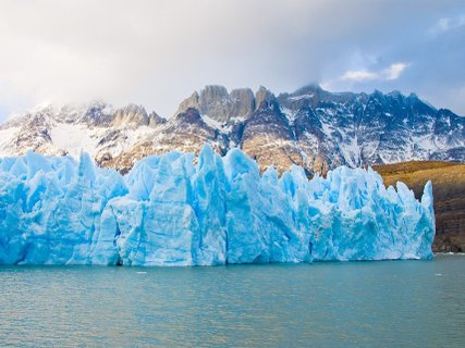 Patagonien Rundreise Selbstfahrer - Grey Gletscher - Gletscherzunge mit zerklüfteter Eisoberfläche vor schroffen, schneebedeckten Bergen und ruhigem Wasser im Vordergrund