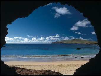 Osterinsel - Blick von einer dunklen Höhle auf einen Sandstrand, das Meer und eine bewachsene Insel unter blauem Himmel mit Wolken