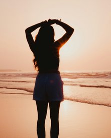 Frau steht bei Sonnenuntergang am Strand und schaut aufs Meer