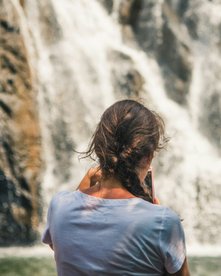 Frau steht vor einem Wasserfall, ihr Rücken zeigt zur Kamera