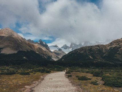 Patagonien Rundreise Selbstfahrer - El Chalten - Wanderweg führt durch karge Vegetation zu bewaldeten Hügeln und wolkenverhangenen Berggipfeln im Hintergrund 