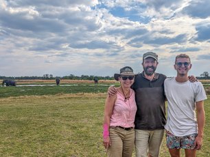 Eine Familie steht lachend vor einer Flusslandschaft in der vereinzelt Elefanten stehen