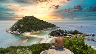 Ein Mann sitzt auf einer Felsanhöhe mit Blick auf eine vorgelagerte tropische Insel