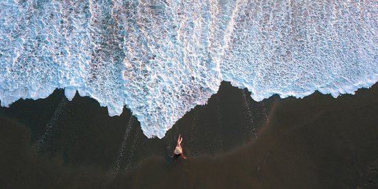 Frau sitzt auf schwarzem Sand vor einer weißen Welle