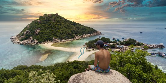 Ein Mann sitzt auf einer Felsanhöhe mit Blick auf eine vorgelagerte tropische Insel