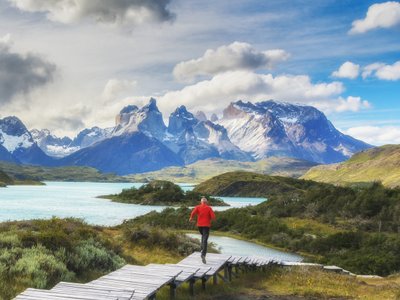 Mensch mit roter Jacke läuft in wunderschöner Landschaft auf einem Steg. Im Hintergrund die Berge des Torres del Paine.