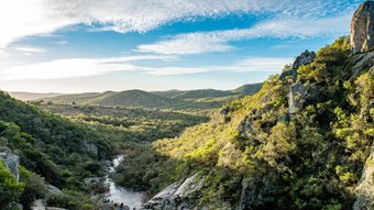 Flusslauf in einem bewaldeten Tal mit Felsen und Hügeln unter blauem Himmel mit Wolken