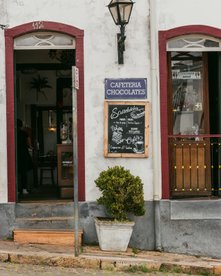Fassade eines Cafés mit offenem Eingang, Schildern mit Texten zu Speisen und Getränken sowie einem Topf mit grünem Busch davor