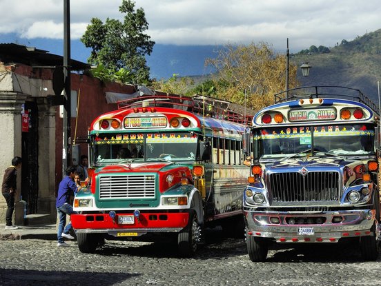 Zwei typisch bunte Busse warten an einer Haltestelle in Guatemala.