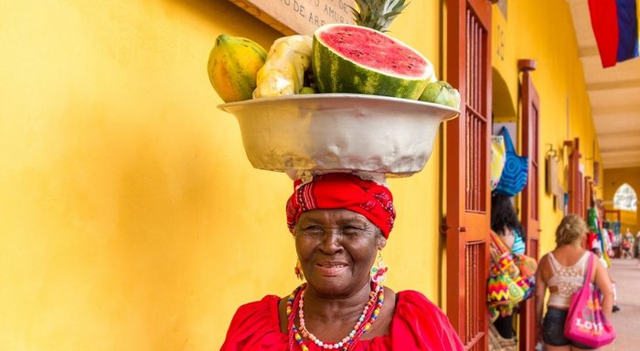 Einheimische Frau mit einer Schale voll Obst auf dem Kopf vor einer gelben Wand