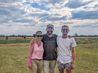 Eine Familie steht lachend vor einer Flusslandschaft in der vereinzelt Elefanten stehen