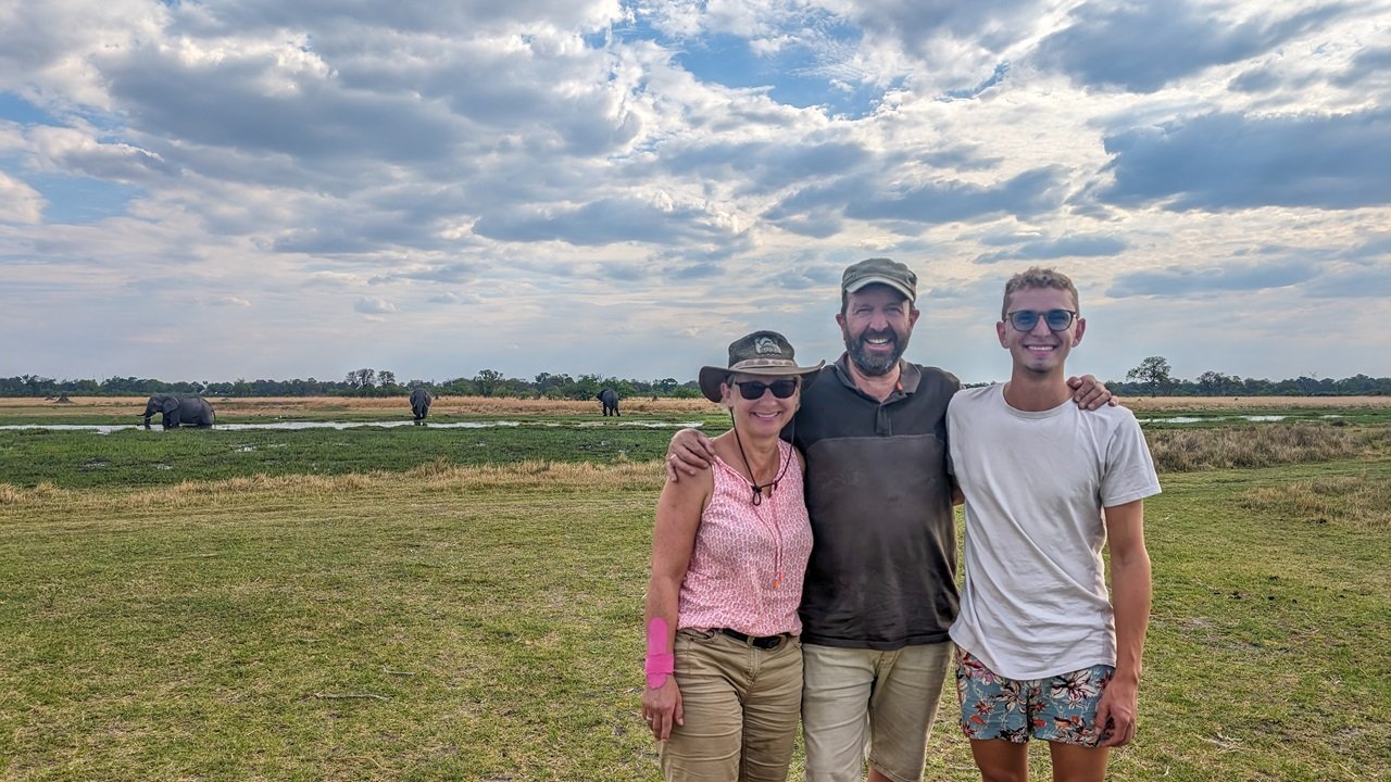 Eine Familie steht lachend vor einer Flusslandschaft in der vereinzelt Elefanten stehen