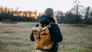 Frau mit gelben Rucksack schaut Sonne zu, wie sie hinter Wald verschwindet