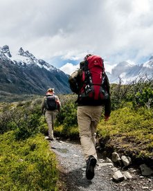 Zwei Wanderer auf einem schmalen Pfad durch grün bewachsene Landschaft mit schneebedeckten Bergen im Hintergrund