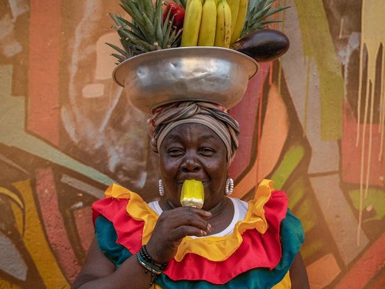 Frau in bunter Kleidung und Obstschale auf dem Kopf schleckt ein Eis