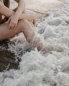 Frau sitzt am Strand und Welle überspült ihre Beine