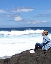 Frau mit Jeansjacke sitzt auf Felsen und schaut aufs Meer