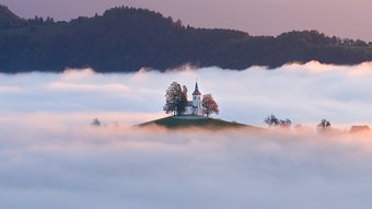 Kirche auf einem Hügel ragt aus Nebelmeer mit Gebirge im Hintergrund beim Sonnenaufgang